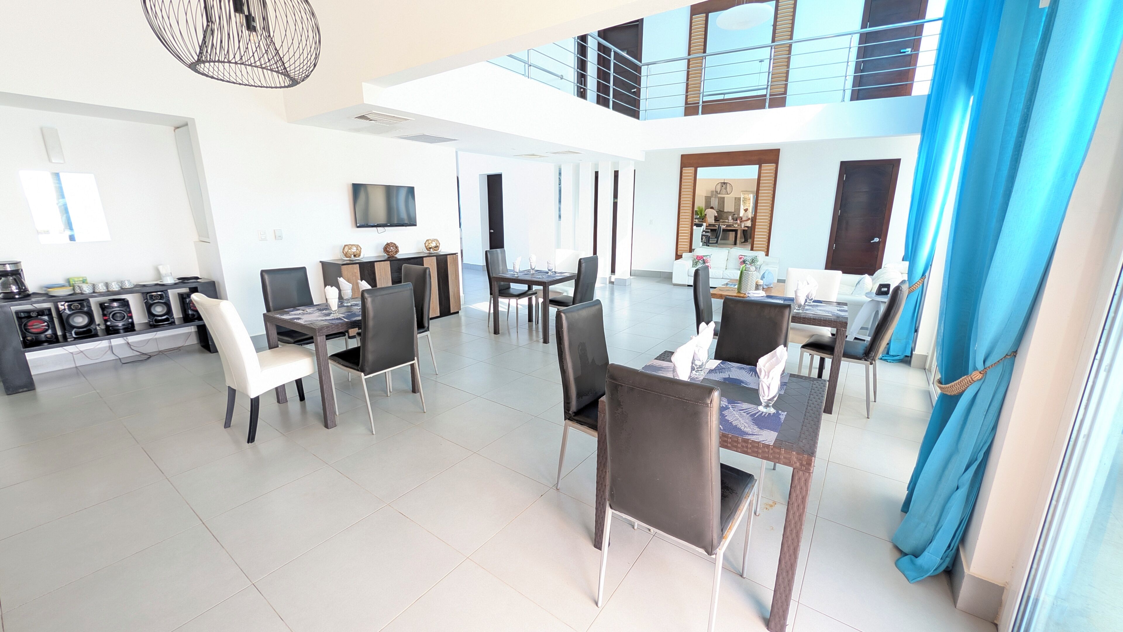 A spacious dining area with multiple tables and chairs, a kitchen area with modern appliances, and a high ceiling with natural light.