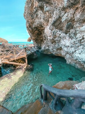 A clear blue water body surrounded by rocky cliffs, a wooden walkway, and a person in a swimsuit. - Peninsula Esmeralda Luxury Hotel & Villas (Puerto Plata)
