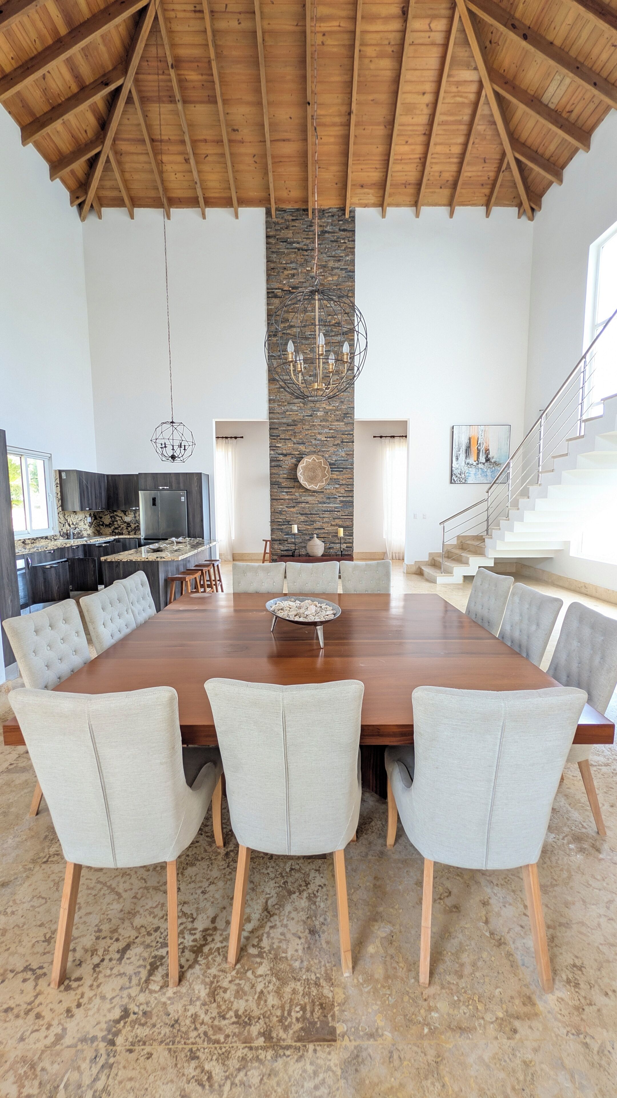 A spacious dining area with a long wooden table and six upholstered chairs, a stone accent wall, and a staircase leading to an upper level.