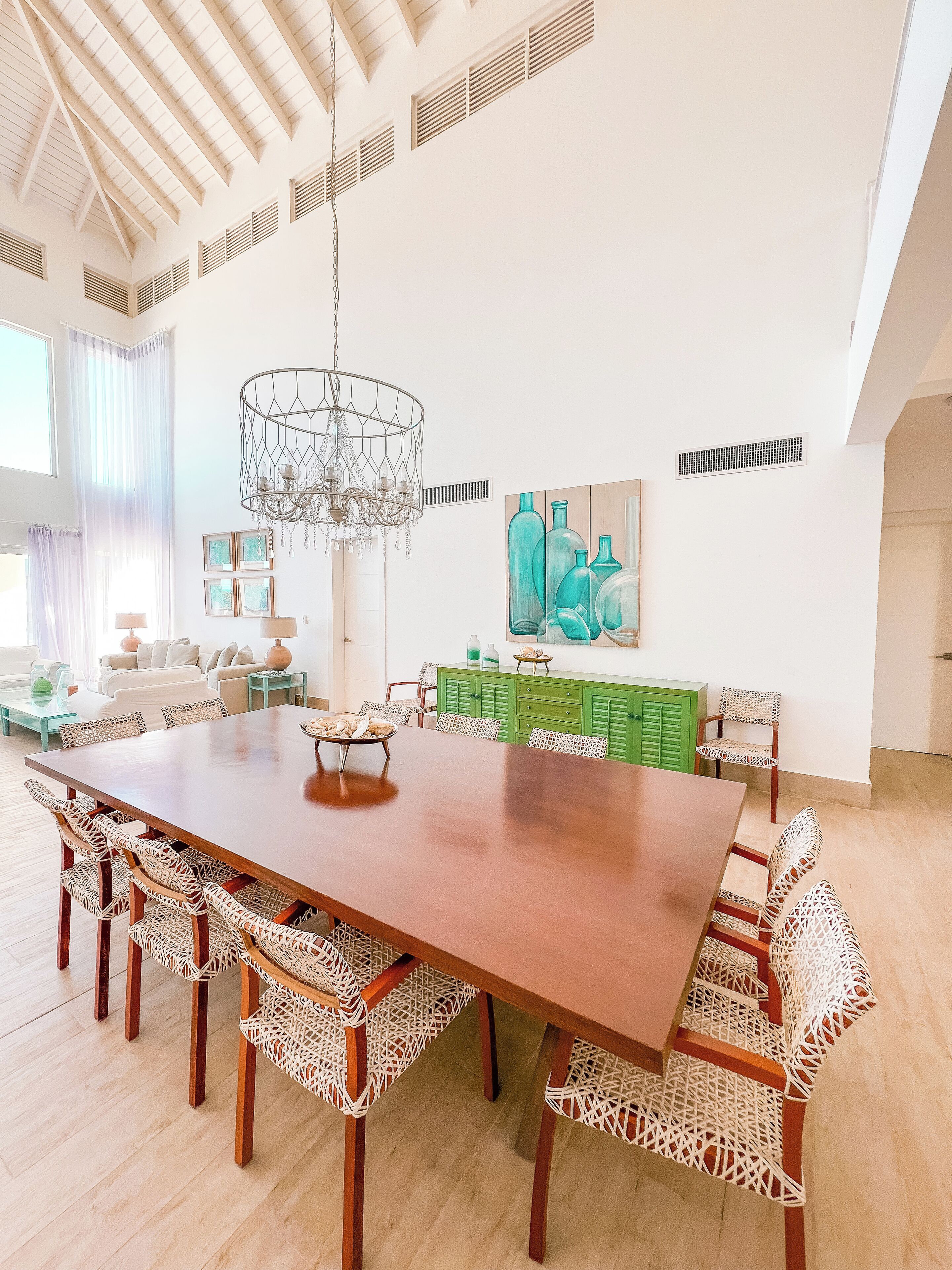 Un comedor amplio con una larga mesa de madera y sillas de mimbre, una araña y un aparador verde con botellas decorativas.