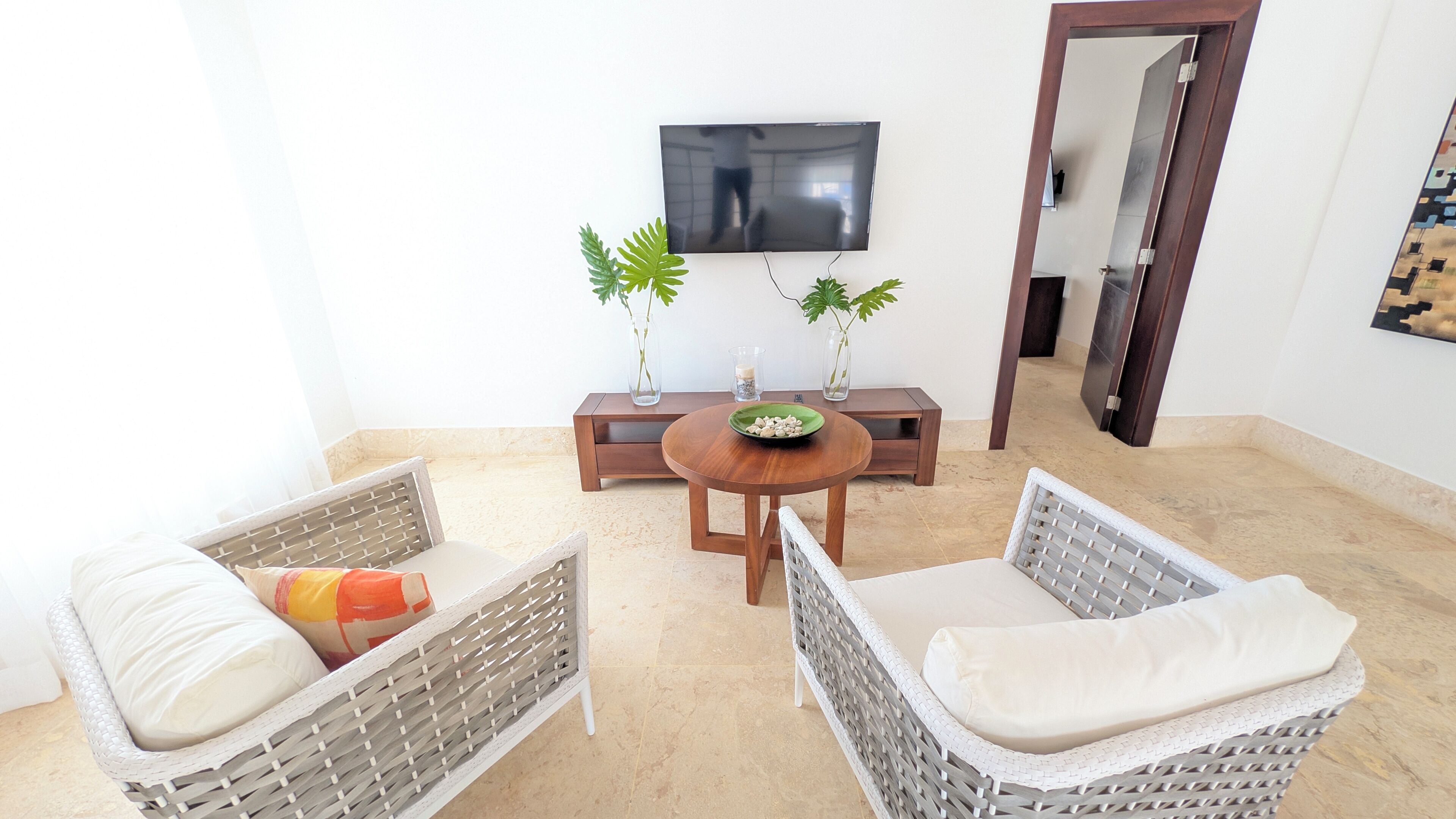 A room with two wicker chairs, a wooden table, a TV mounted on the wall, and two potted plants.