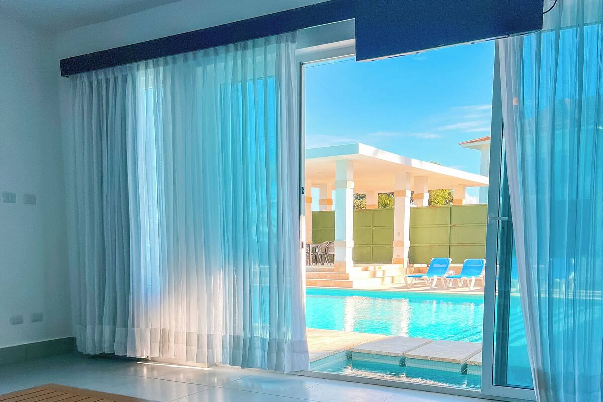 Ein Hotelzimmer mit Bett, Blick auf einen Pool und großen Fenstern mit Vorhängen.