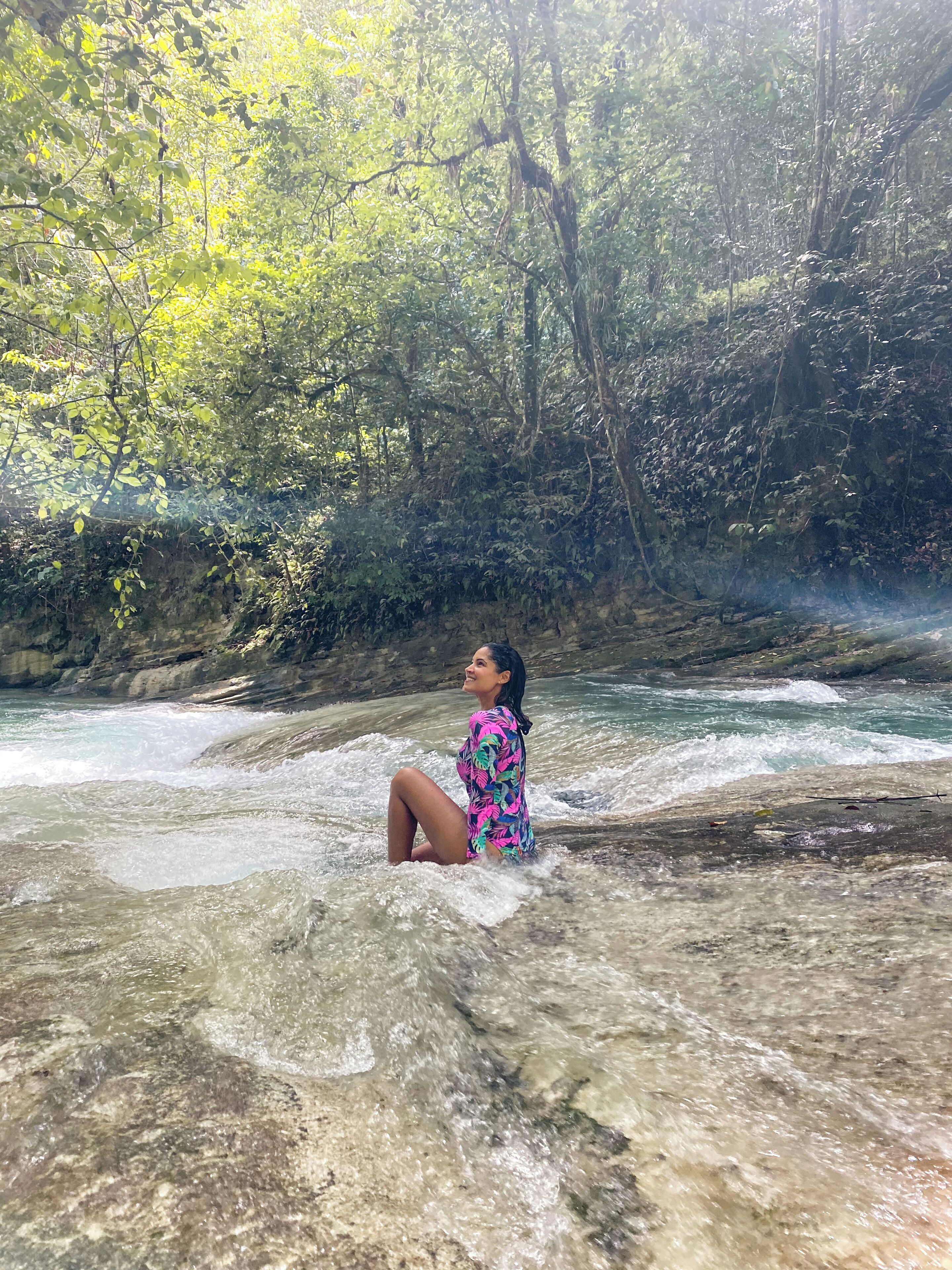 A person sitting on a rocky surface by a flowing river, surrounded by dense greenery.