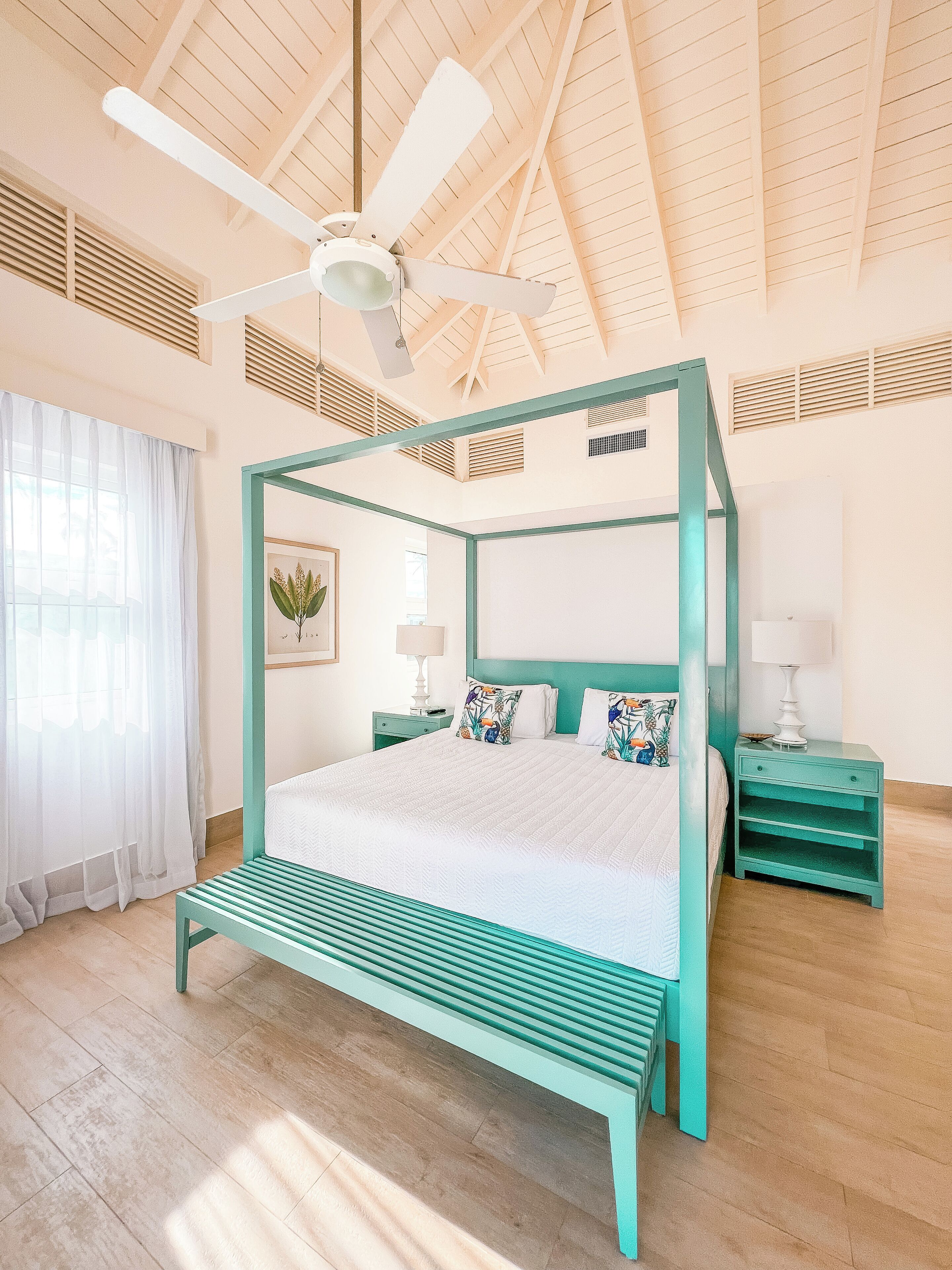 Un lit à baldaquin avec une couvre-lit blanche et des touches de couleur turquoise, un ventilateur de plafond et une fenêtre ornée de rideaux légers.