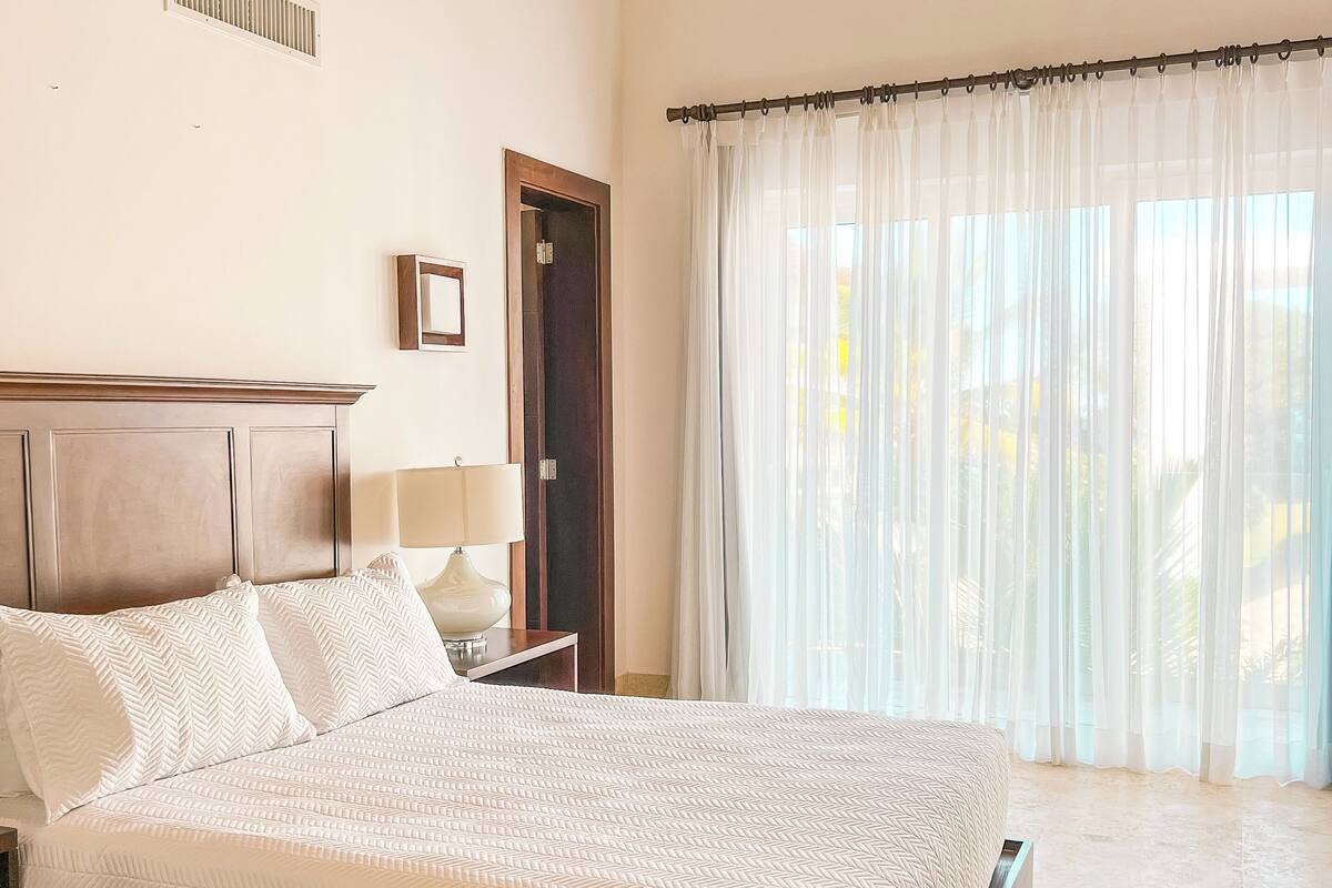 A bedroom with a bed, a ceiling fan, a nightstand, a lamp, and a window with sheer curtains.