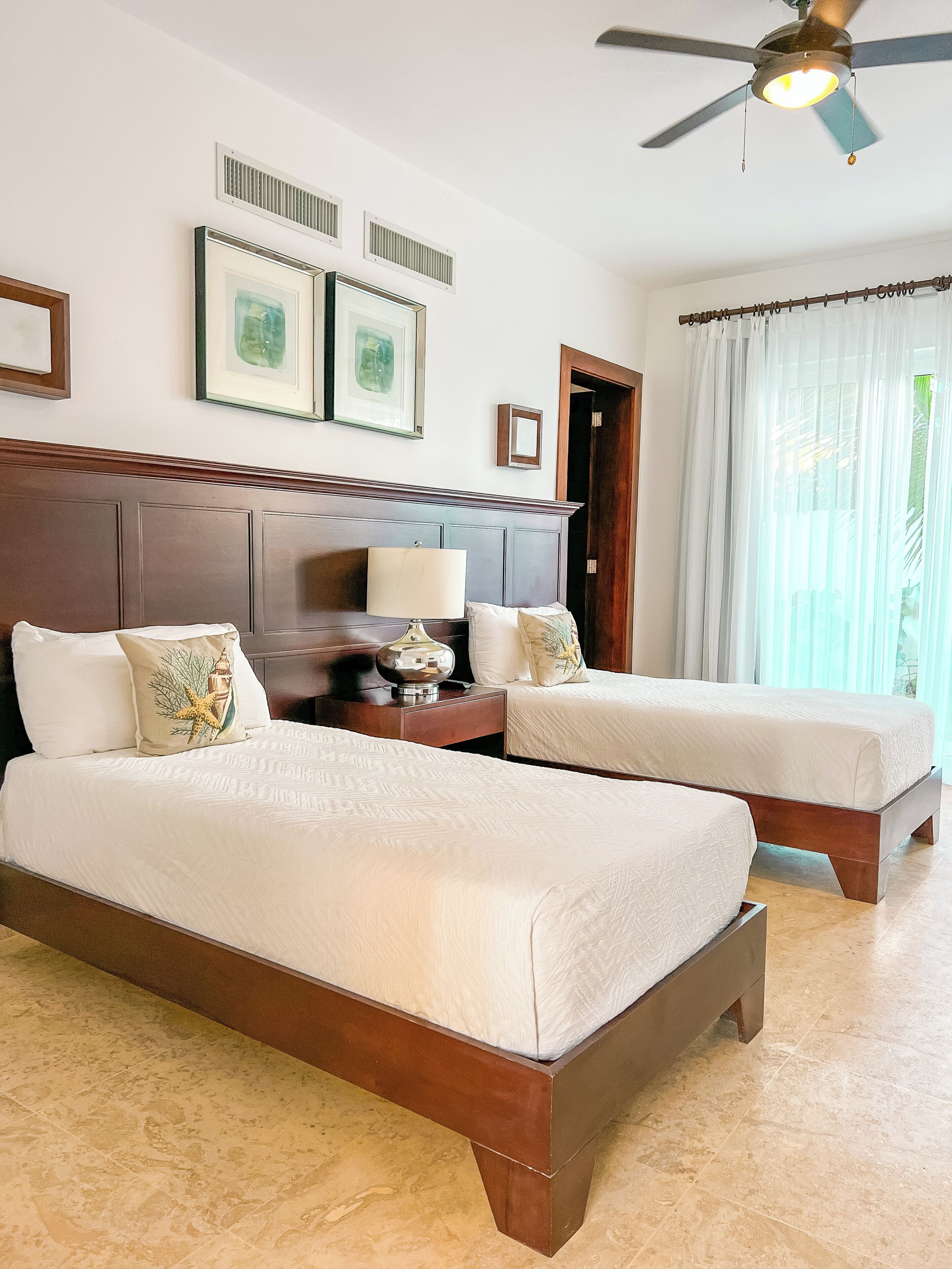 A hotel room with two beds, a ceiling fan, framed artwork, and a window with sheer curtains.