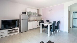 Een moderne keuken met witte kasten, een ingebouwde magnetron en een roestvrijstalen koelkast. Er is een eettafel met vier stoelen en een flatscreen televisie op een standaard.