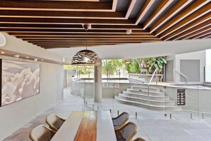Ein modernes Interieur mit einem langen Esstisch, Korbstühlen, einer Holzbalkendecke und einer Treppe, die zu einem Außenbereich führt.