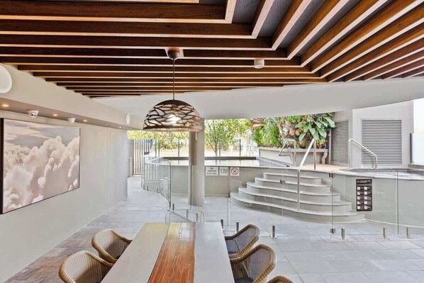 Un intérieur moderne avec une longue table à manger, des chaises en osier, un plafond avec des poutres en bois et un escalier menant à un espace extérieur.