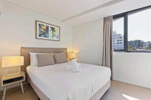 Une chambre d’hôtel avec un grand lit, une table de chevet avec une lampe, un tableau accroché au mur et une vue sur les immeubles par la fenêtre.