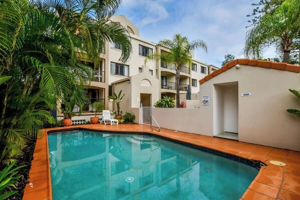 Une piscine dont le bord est recouvert de carreaux, entourée de palmiers et bordée d’un bâtiment doté de balcons.