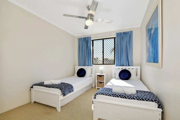 Habitación doble con dos camas, ventilador de techo, ventana con cortinas y una mesita de noche pequeña.