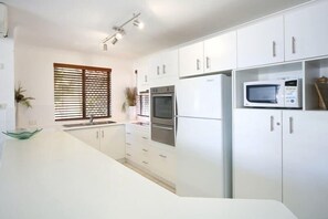 A modern kitchen with white cabinetry, a built-in oven, a microwave, and a window with blinds. - Beachport 16 Waterfront Views Apt (Mooloolaba)