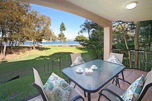 A balcony with a glass table and chairs, overlooking a grassy area and a body of water. - Beachport 16 Waterfront Views Apt (Mooloolaba)