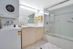 A modern bathroom with a built-in washing machine, a glass shower enclosure, and a large mirror. - Lamer 5 Beachfront Apt Mooloolaba (Maroochydore)