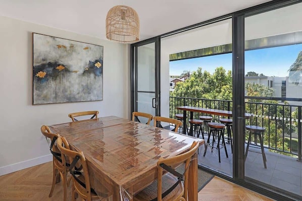 Un coin repas avec une table et des chaises en bois, un grand tableau au mur et un balcon avec des sièges d’extérieur.