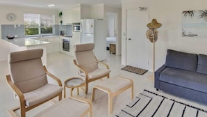 Uma sala de estar moderna com um sofá, duas poltronas e uma mesa de centro. Há um ventilador de teto, um relógio e uma janela com persianas.