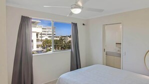Quarto com cama, ventilador de teto, janela com cortinas e vista para edifícios.