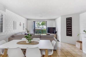A modern living room with a white sofa, dining table, and chairs. There is a TV on a stand, a large window, and a wall-mounted sign listing beach names.