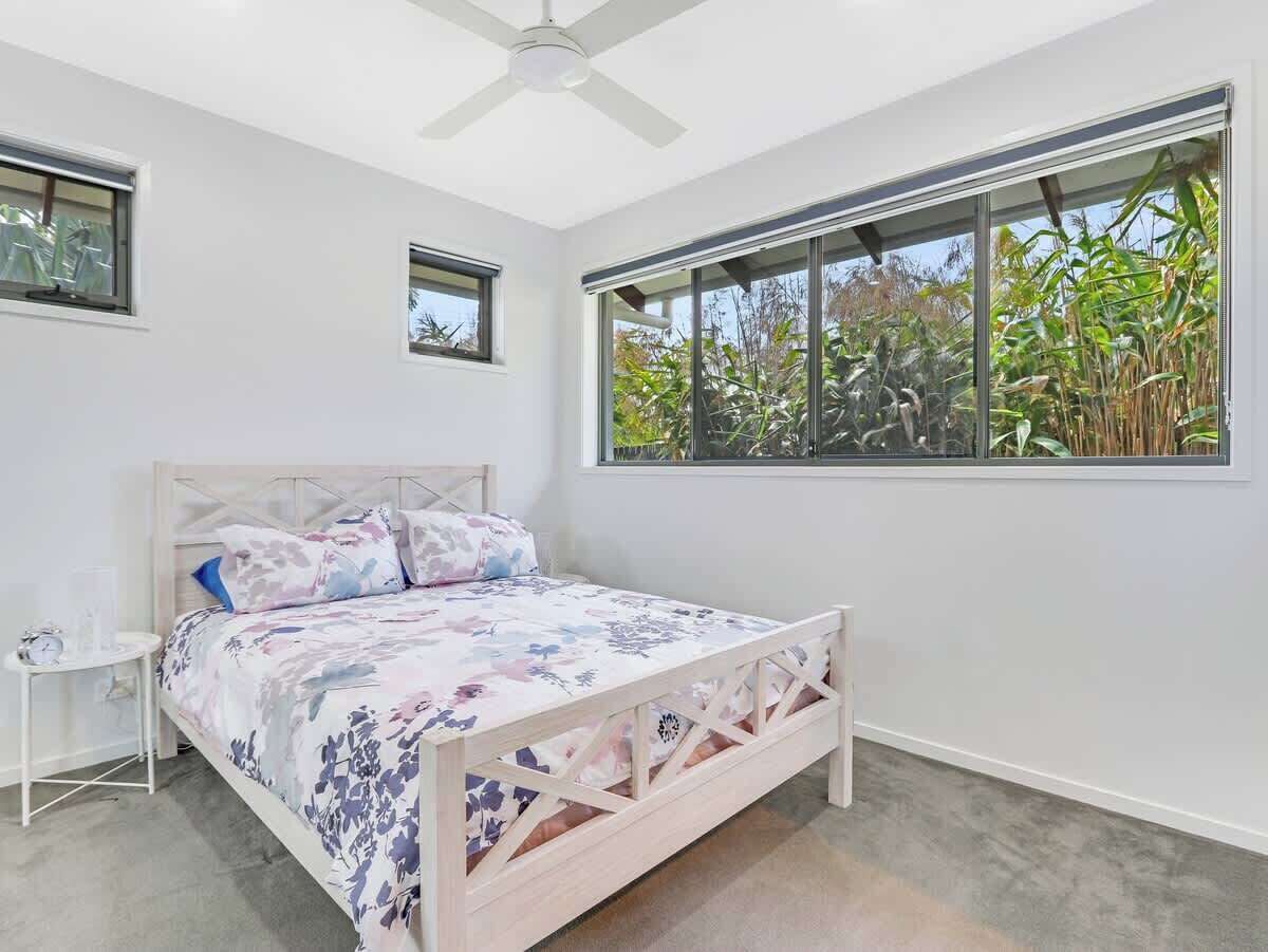 A bedroom with a bed, a nightstand, a ceiling fan, and a large window with a view of greenery.