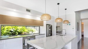 A modern kitchen with a large island, stainless steel appliances, and a view of greenery outside. - Adelong Waterfront Home Mooloolaba (Buddina)