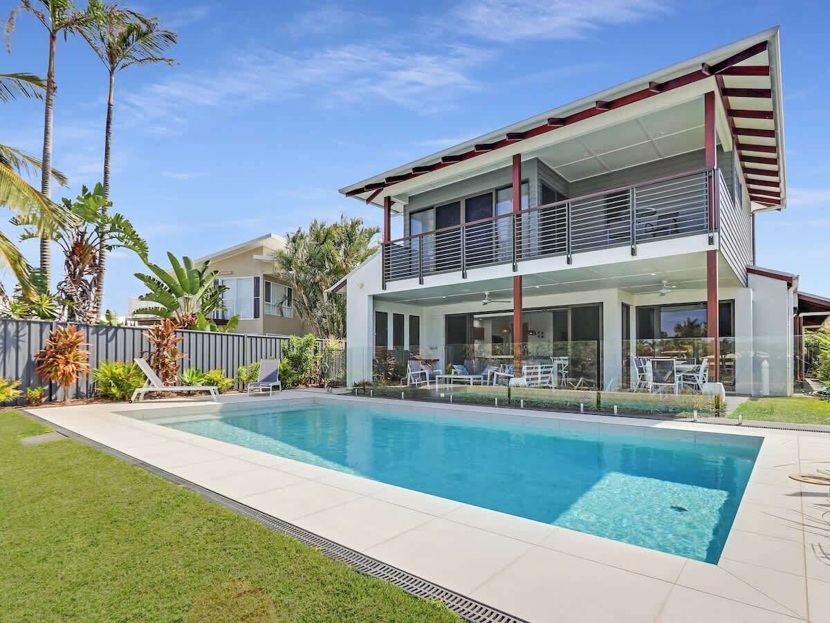 Una casa moderna de dos pisos con piscina, rodeada de plantas tropicales y un césped bien cuidado.