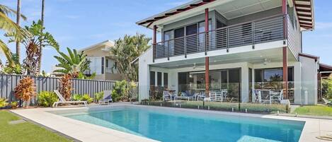 Una casa moderna de dos pisos con piscina, rodeada de plantas tropicales y un césped bien cuidado.