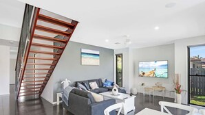 A modern living room with a grey sofa, a white coffee table, and a dining area with white chairs. A wooden staircase leads to an upper level. - Adelong Waterfront Home Mooloolaba (Buddina)