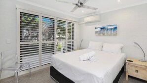 A bedroom with a bed, a ceiling fan, a nightstand, a chair, and a window with blinds. - Illawong 1 Beachfront Doorstep Apt (Mooloolaba)