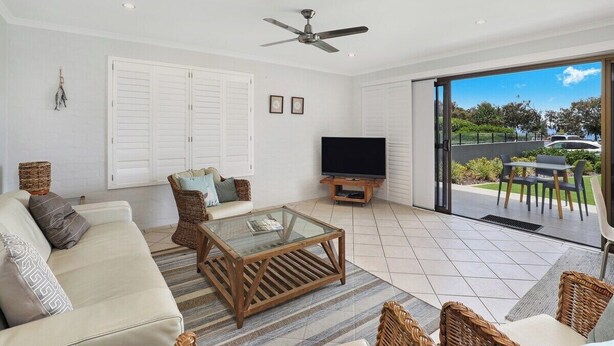 A modern living room with a sofa, coffee table, and TV, connected to an outdoor patio with a table and chairs. - Illawong 1 Beachfront Doorstep Apt (Mooloolaba)