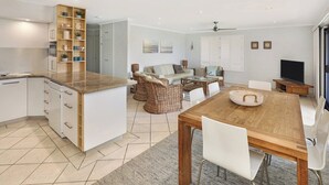 A modern kitchen with white cabinets, a wooden island, and a dining area with a wooden table and white chairs. - Illawong 1 Beachfront Doorstep Apt (Mooloolaba)
