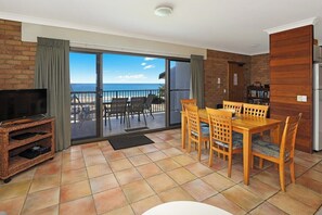 A room with a dining table and chairs, a television, and a view of the beach through a sliding glass door.
