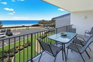 Een balkon met een glazen tafel en stoelen, met uitzicht op een parkeerplaats en een strand.