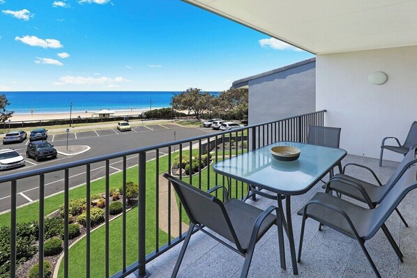 A balcony with a glass table and chairs, overlooking a parking area and a beach.