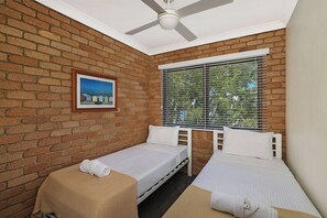 A room with two single beds, a ceiling fan, a window with blinds, and a framed picture on the brick wall.