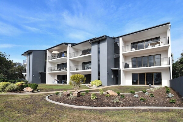 A modern multi-story building with balconies, a landscaped garden, and a clear blue sky.
