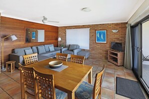 A cozy living area with a wooden dining table, grey sofas, and a TV on a brick wall.