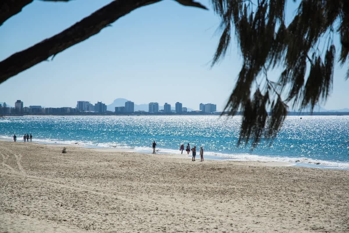 A beach with people walking along the shore, a clear sky, and a city skyline in the background.