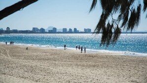 Ein Strand mit Menschen, die am Ufer entlanggehen, einem klaren Himmel und einer Stadtansicht im Hintergrund.