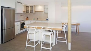 Une cuisine moderne avec des meubles blancs, un réfrigérateur en acier inoxydable, un îlot central doté de tabourets de bar et une chaise en osier.