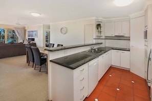 Cocina compacta con armarios blancos, encimera negra y zona de comedor con sillas.