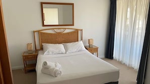 Une chambre d’hôtel avec un grand lit, une tête de lit en bois, deux tables de chevet avec des lampes, un miroir au-dessus du lit et une fenêtre avec des rideaux.