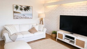 Un salon avec un mur en briques blanches, un canapé blanc, un téléviseur à écran plat et un tableau représentant une plage.