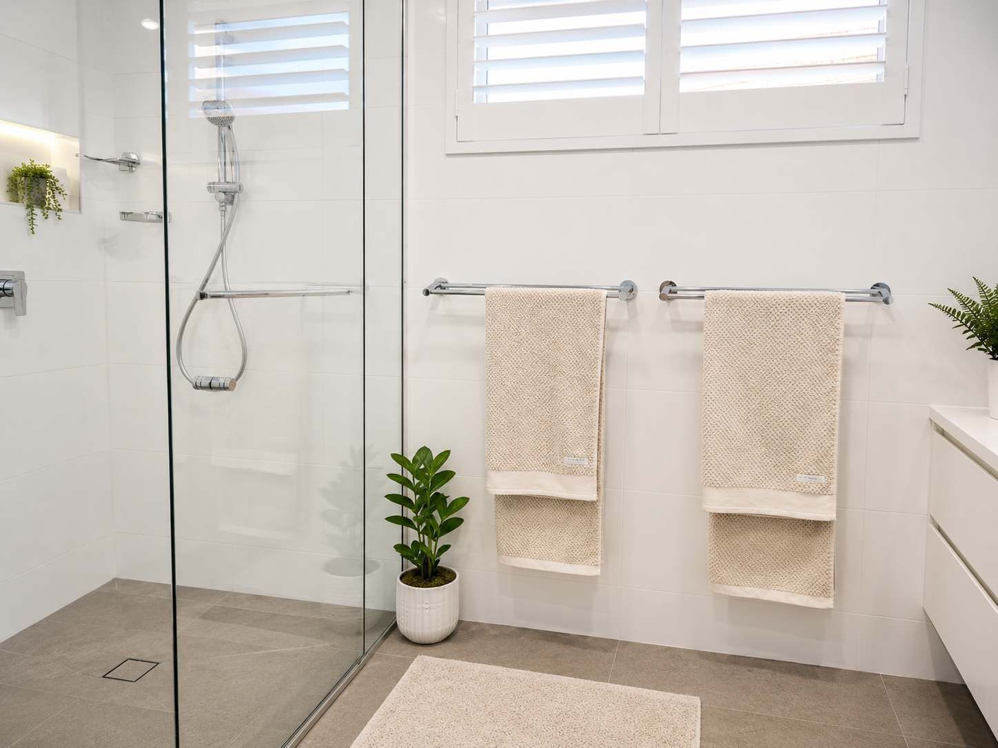 A modern bathroom with a glass shower enclosure, two towels on a rack, a potted plant, and a window with blinds.