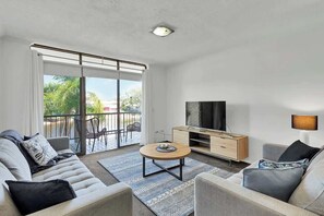 Una sala de estar moderna con sofá, mesa de centro, televisor y un balcón con mobiliario de exterior.