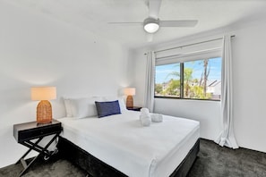A hotel room with a bed, two bedside tables, a ceiling fan, a window with curtains, and a view of buildings outside. - Hacienda 3 Canal View Comfort Apt (Mooloolaba)