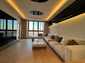 A modern living room with a large sectional sofa, a coffee table, and a view of the city through large windows. - Luxury Chic Suite In Aeon Compound- Mall Of Arabia Towers (Giza Governorate)