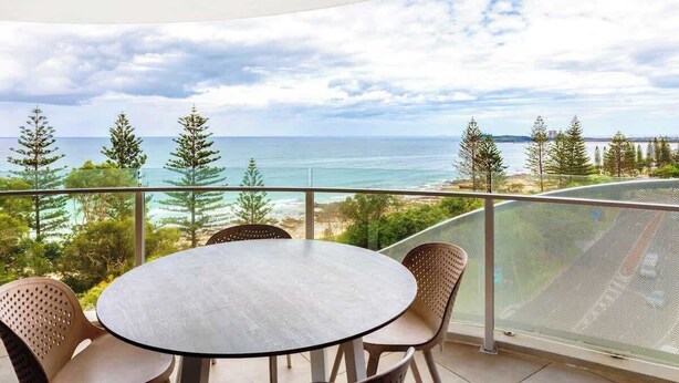 A balcony with a round table and chairs, overlooking a coastal landscape with trees and the sea. - Breeze 801 Luxury Mooloolaba Apt with Ocean Views (Mooloolaba)