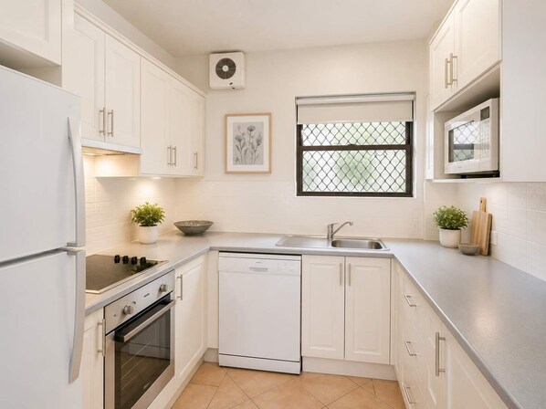 A modern kitchen with white cabinetry, a dishwasher, an oven, a microwave, a sink, and a window with a metal screen. - Warroo 5 Pool Apt Alex Headland (Alexandra Headland)