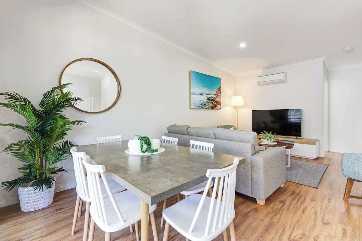 A modern living room with a grey sofa, a flat-screen TV, a dining table with white chairs, and a large potted plant.